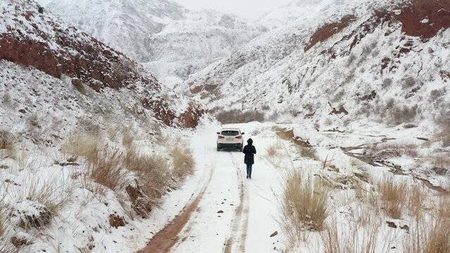 A girl in a black hooded jacket walks along a snowy mountain road to a white SUV and takes pictures of everything around. Travel to Kyrgyzstan. Beautiful landscape.