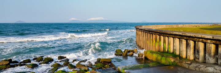 Seascape with moss-covered rocks and concrete sea wall against waves, with distant islands on the horizon, ideal for travel and nature themes with copy space on the right - obrazy, fototapety, plakaty
