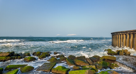 Seascape with moss-covered rocks and waves crashing against a concrete breakwater, with clear sky...