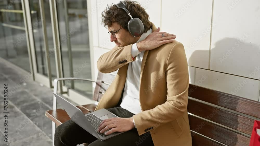 Poster A stylish man in a beige coat works on his laptop with headphones on a bench in an urban setting