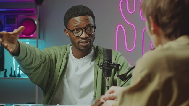 Young African American blogger speaking in microphone as recording podcast with female cohost in modern studio with trendy neon decor. Over-the-shoulder view