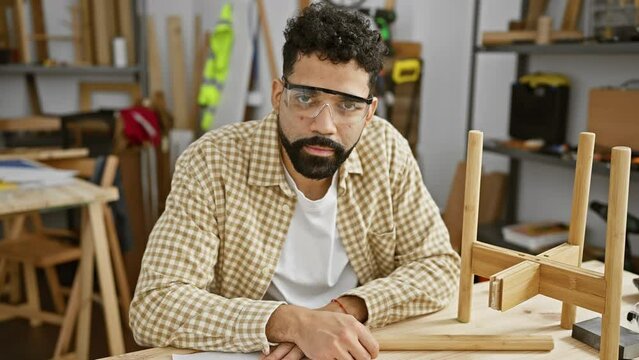 Handsome hispanic man with beard wearing safety glasses in a carpentry workshop looking at camera.