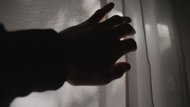 Hand reaching towards white window curtain - opening fabric to let sun shine inside dark room. Flare hitting camera. Green backyard outside house or apartment window