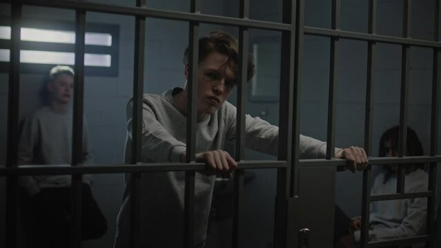 Multicultural teenage prisoners behind metal bars in prison cell look at camera. Young criminals serve imprisonment terms for crimes in jail. Youth detention center or correctional facility. Portrait.