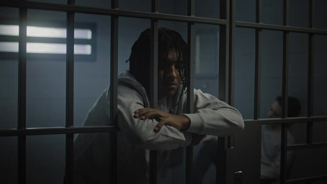 Depressed African American teenager with tattoos stands in prison cell in jail and looks at camera. Young inmate serves imprisonment term for crime. Juvenile detention center or correctional facility.