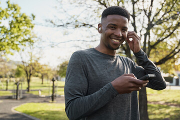 Young black man in sports clothing with wireless earphones and mobile phone smiling in park - 762954441