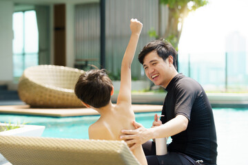 Father is applying a sun screen or sun block lotion on his son body before going to swimming in the swimming pool. 