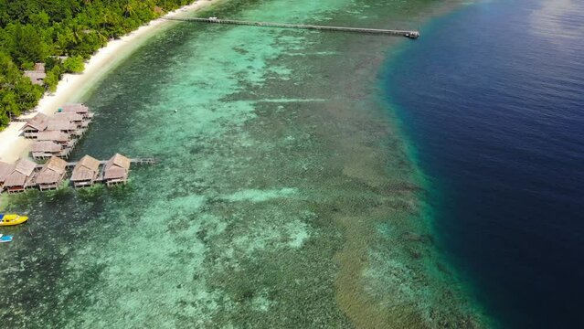 Clear turquoise and blue water in front of sandy beach with wooden walkway, pier and wooden guesthouses, Lumba Lumba guesthouse on Kri Island in Raja Ampat, Indonesia .