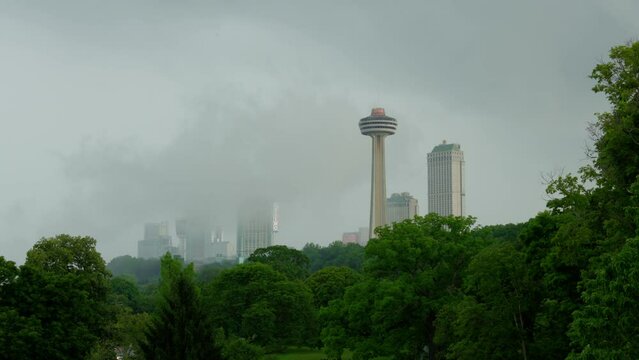 Picturesque shot of the Skylon Tower in Niagara Falls, Ontario, with mist rising from The Falls, framed by a canopy of green, lush trees.
