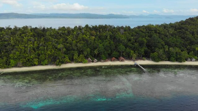 Perfectly clear water showing turquoise ocean floor in front of island paradise of Kri in Raja Ampat, Indonesia with palm trees and vacation huts, aerial dolly out.