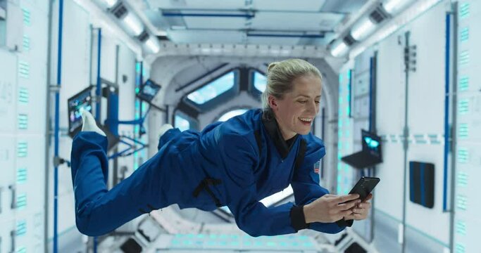Cheerful Female Astronaut Floating in Zero Gravity Inside a Spaceship. Happy Specialist Using Smartphone in Space, Checking News and Communicating with Family and Friends Online 