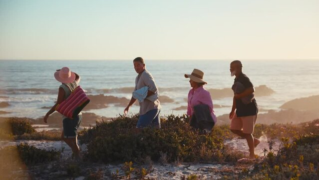 Camera tracks group of mature couples having fun on summer vacation walking through dunes on way to beach carrying bags and towels - shot in slow motion