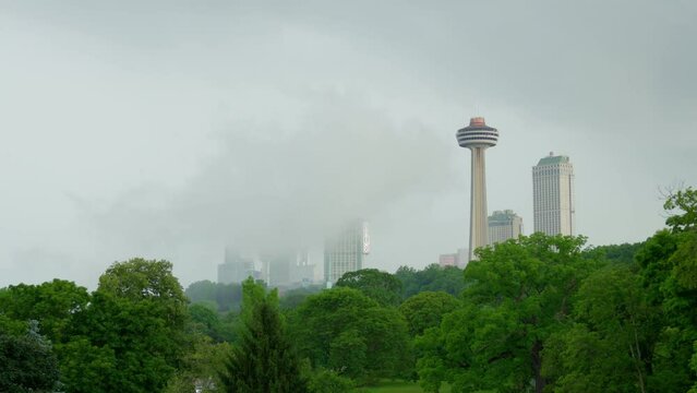 The camera pans to the right as we see mist that has risen up from Niagara Falls, partially obscuring the Niagara Falls skyline. Skylon Tower stands tall, bursting forth out of lush greenery.