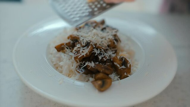 Woman's hands grating parmesan cheese over the risotto with sliced mushrooms. Close up