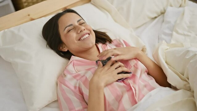 Hispanic woman smiling while using smartphone in bedroom, depicting leisure and comfort at home