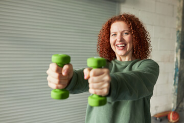 Selective focus on red head female with curls outstretching hands with green dumbbells forward,...