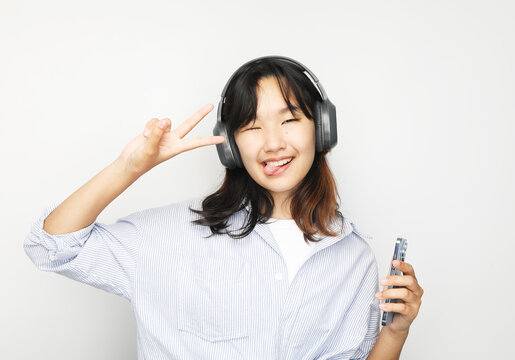 Cheerful Asian teenage girl listens to music on headphones and shows a victory sign.