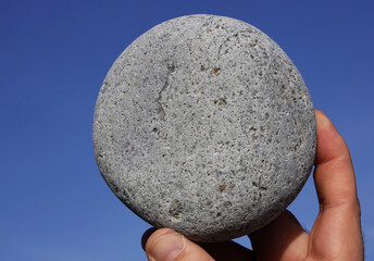 round pebble stone hold in male hand on blue sky background.
