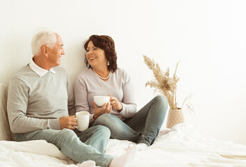 Happy elderly couple drinking coffee sitting on bed on bedroom.