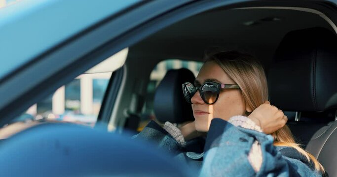 attractive blonde woman sitting in car and looking in sun visor vanity mirror to arrange hair and sunglasses