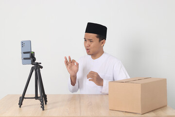 Portrait of excited Asian muslim man in koko shirt with skullcap promoting his product on live...