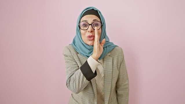 Hispanic middle-aged woman spills secrets over isolated pink background, whispering malicious tales, hijab concealing her mouth, hand mid-gesture during engrossing conversation