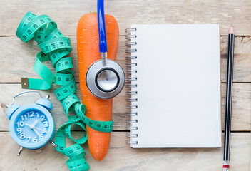 Top view carrot with blue stethoscope,measuring tape,alarm clock and notebook on wood table...