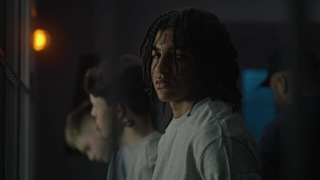 African American teenager stands near prison cell with Caucasian boys, looks at the camera. Young prisoner serves imprisonment term for crime in jail. Youth detention center or correctional facility.