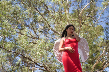 South American woman, young, beautiful, brunette with red dress and white wings, posing in the middle of nature, pure, alone, relaxed and calm. Concept angels, beauty, costumes, wings, halloween.