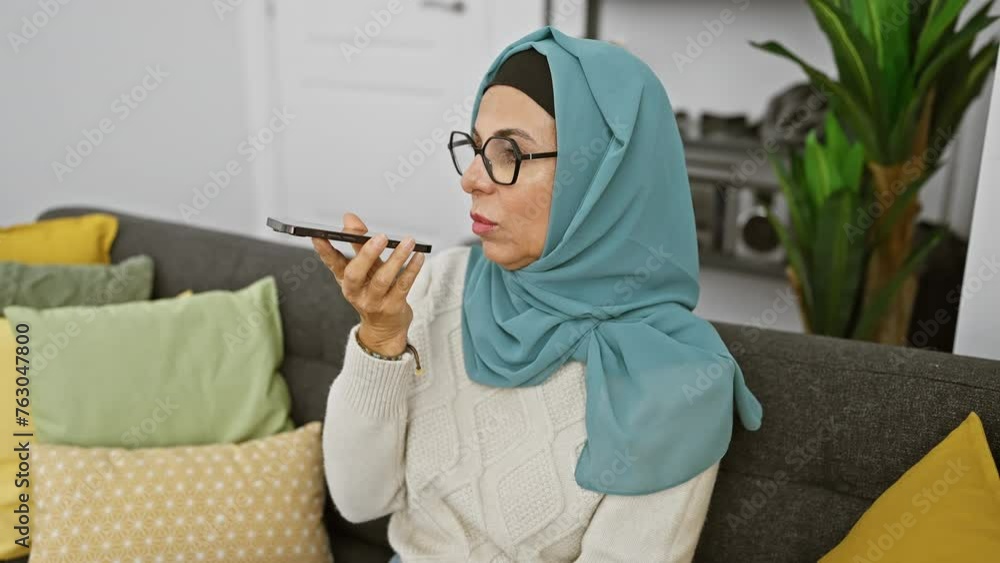 Canvas Prints a mature woman wearing a hijab sends a voice message on her smartphone in a cozy living room.