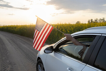 white car with an American flag hanging from the window drives along a country road among a...