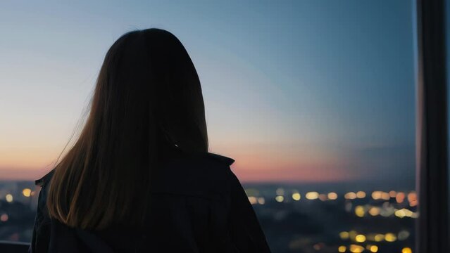 Woman's back turned as she stares out at cityscape during twilight. Stands on rooftop of tall building, taking in view of neon-drenched streets below. 