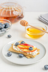 Healthy sweet sandwiches made of crispbread thins and cream cheese decorated with sliced nectarine fruit, blueberries and flax seeds served on plate on white wooden table with glass tea pot and honey