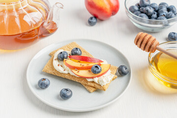 Healthy homemade sweet sandwiches made of crispbread and mascarpone cheese decorated with sliced nectarine fruit, blueberries and flax seeds served on plate on white table with tea pot and honey