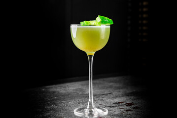 A refreshing cocktail with lime garnish in a stemmed glass, showcased against a dark, moody background. The drink features a yellowish-green hue, possibly indicating it’s a citrus or herbal cocktail.