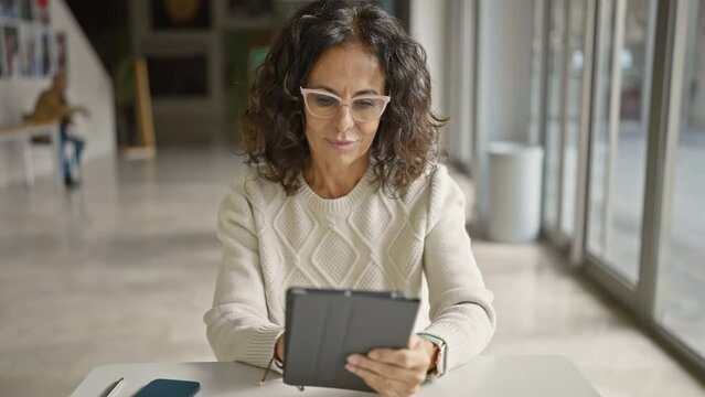 A professional hispanic woman wearing glasses focused on her tablet in a modern office setting.