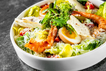 A fresh Caesar salad with grilled shrimp, greens, egg, croutons, and cherry tomatoes in a white bowl on a grey surface. The salad is topped with grated cheese and fresh herbs