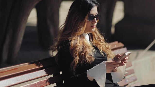 Elegant, business woman on a break in the park writes a message with a phone in her hands, remote work, communication on social networks, business tone, mobile phone, style and restraint, technology