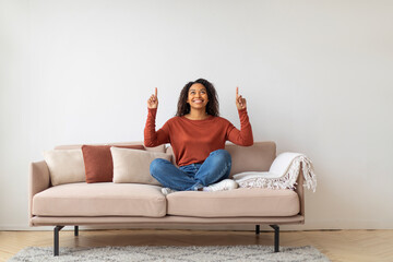 Cheerful young black woman sitting cross-legged on sofa, smiling and pointing upwards