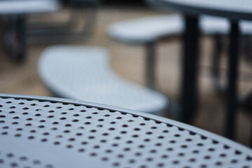 Closeup of the edge of a park table. The steel design has holes cut out to allow water to pass through. In the background, defocused benches are visible.
