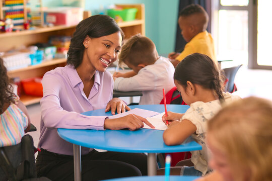 Female Primary Or Elementary School Teacher Helping Students At Desks In Multi-Cultural Class 