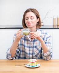 Close-up portrait of attractive girl drinking hot matcha tea and eating tasty cake in the kitchen	