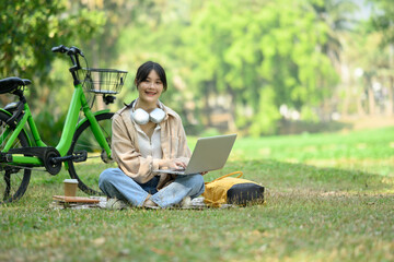 Full body shot of smiling Asian woman sitting on grass near bicycle and using laptop