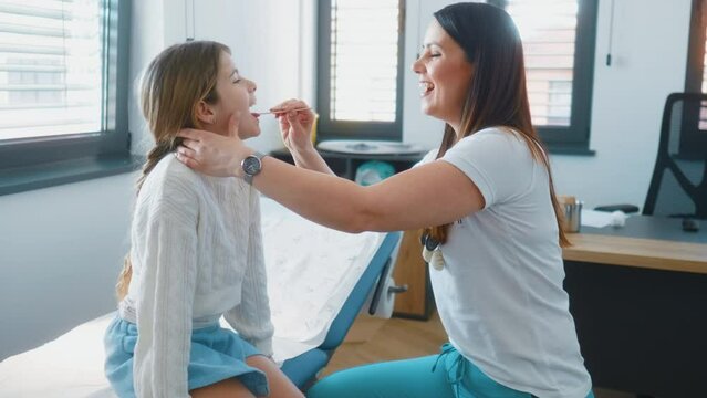 Female pediatrician with tongue depressor checking little girl throat on medical exam at clinic