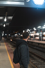 Young man facing away in black and grey jacket and black backpack waits for his train on a plataform in Vigo Guixar railway station on a winter night.