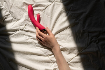 female hand reaching for a pink sex toy rabbit shaped vibrator for women in bed, sun rays on a quilted white blanket, good morning concept