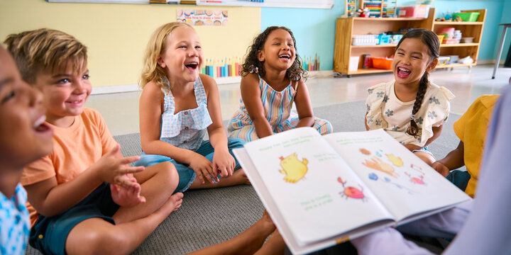 Female Primary Or Elementary School Teacher Reads Story To Multi-Cultural Class Seated In Classroom
