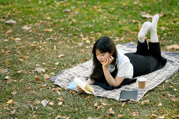 Full body photo of young cute asian woman lying on blanket and reading book in the park