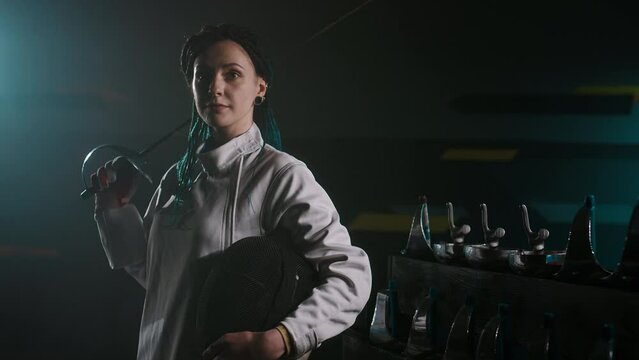 Medium portrait of young female foil fencer in padded uniform posing with foil sword and epee mask and looking at camera while standing in dark room