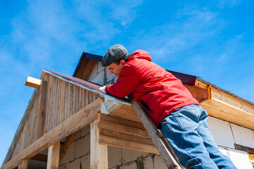 A worker builds a roof in a house while standing on a wooden ladder. Blue sky